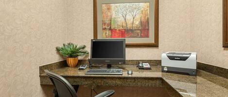A hotel reception desk with a desktop computer, a framed painting, a plant, a telephone, and a printer.