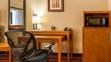A hotel room with a wooden desk, a black office chair, a microwave, a telephone, a lamp, and a framed picture on the wall.