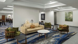 A modern lobby with a beige sofa, green armchairs, and a round coffee table. There is a large abstract rug, a painting of a forest, and a sign that reads "LESS MONDAY MORE SUMMER". - Fairfield Inn & Suites South Boston (South Boston)