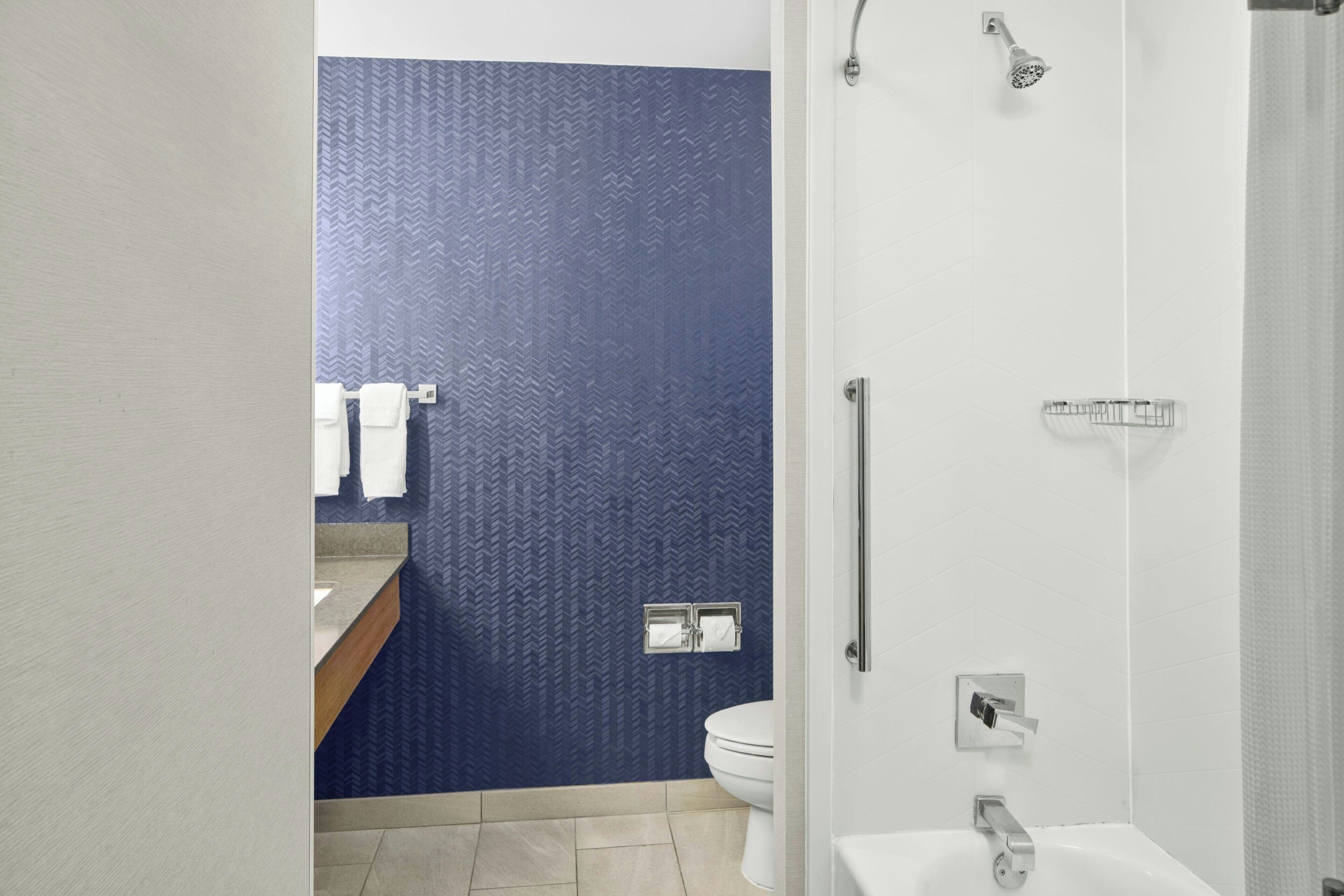 Un baño moderno con una pared azul texturizada, un inodoro blanco, una ducha con cabezal de ducha manual y un lavamanos con toallero.