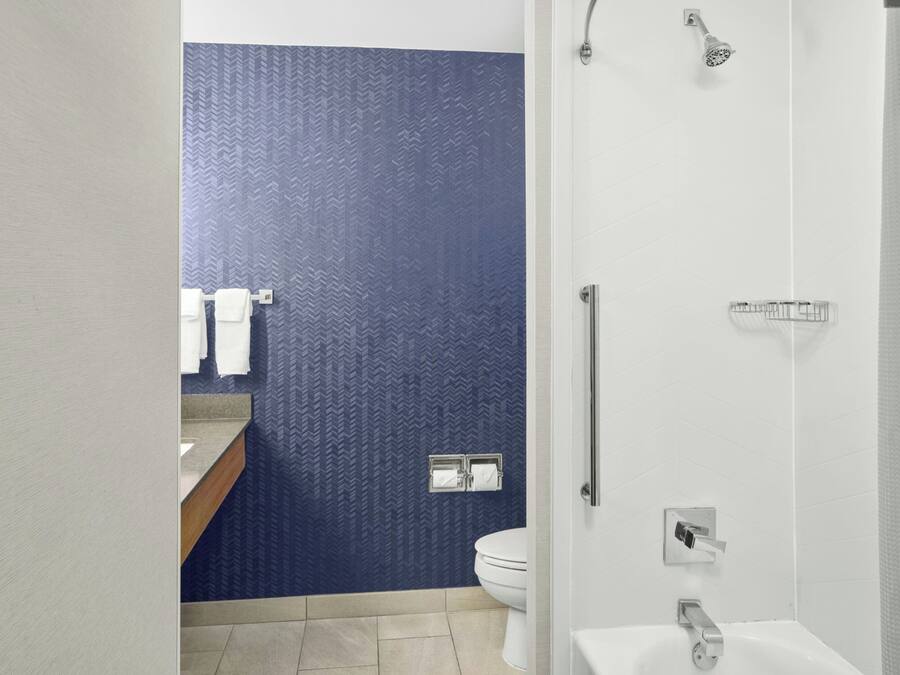 Un baño moderno con una pared azul texturizada, un inodoro blanco, una ducha con cabezal de ducha manual y un lavamanos con toallero.