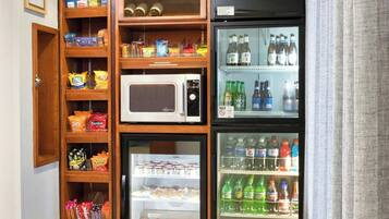 A small kitchenette with a microwave, refrigerator, and shelves stocked with snacks and beverages.