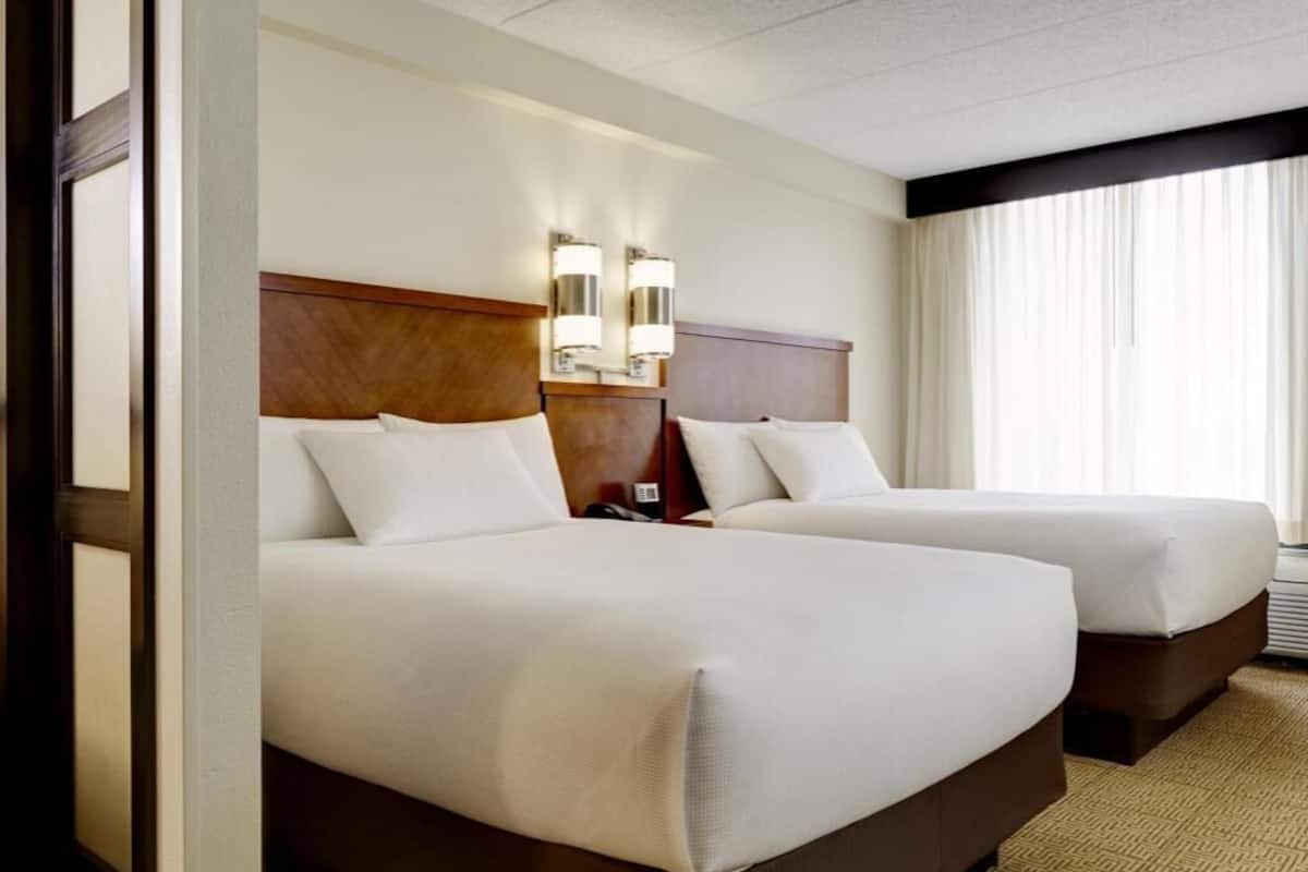 Ein Hotelzimmer mit zwei Betten, einem Holzkopfstück, Wandlampen und einem großen Fenster mit Vorhängen.