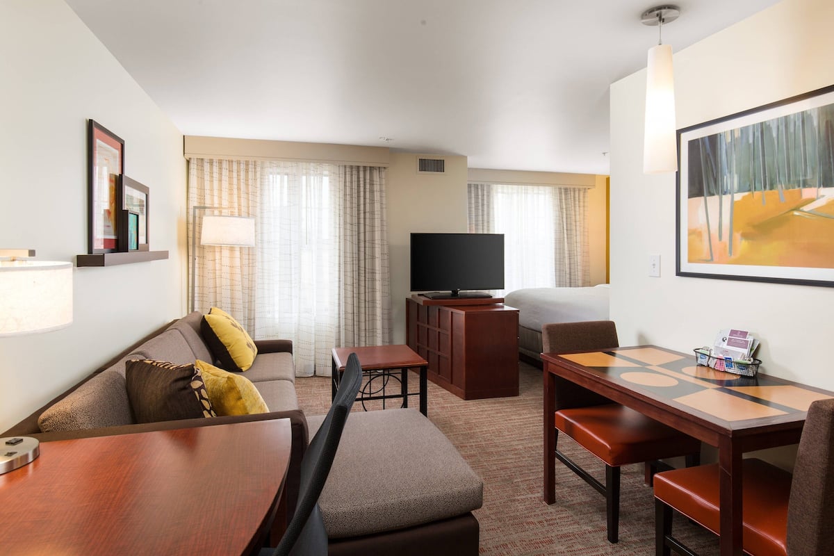 A hotel room with a sofa, a TV, a dining table, and a large painting on the wall.