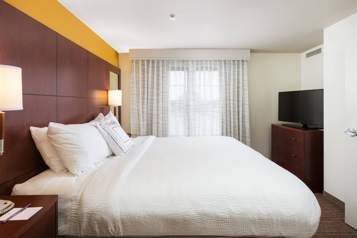 A hotel room with a large bed, a wooden headboard, a television on a wooden cabinet, and a window with curtains.