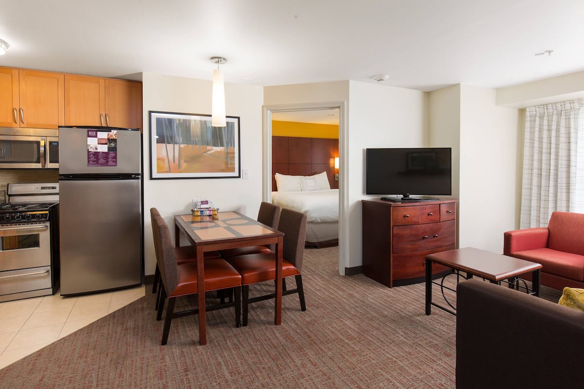 A compact hotel room with a kitchenette, a dining area, a bed, a TV, and a red sofa.