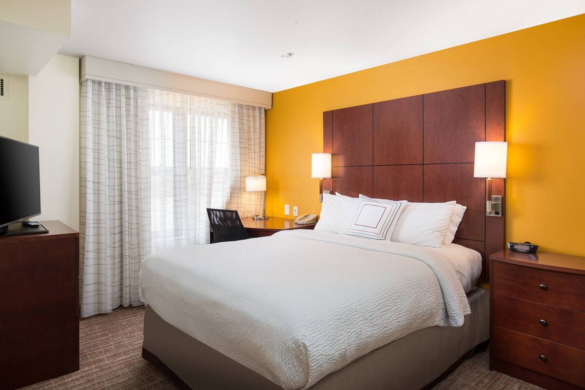 A hotel room with a large bed, two bedside lamps, a desk with a chair, and a television on a stand.