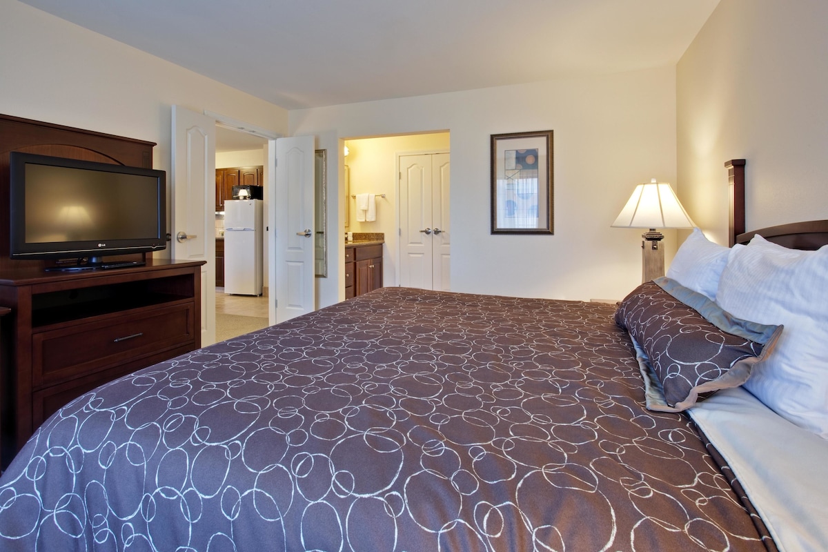 A hotel room with a large bed, a TV on a wooden stand, a lamp, and a door leading to a kitchen.