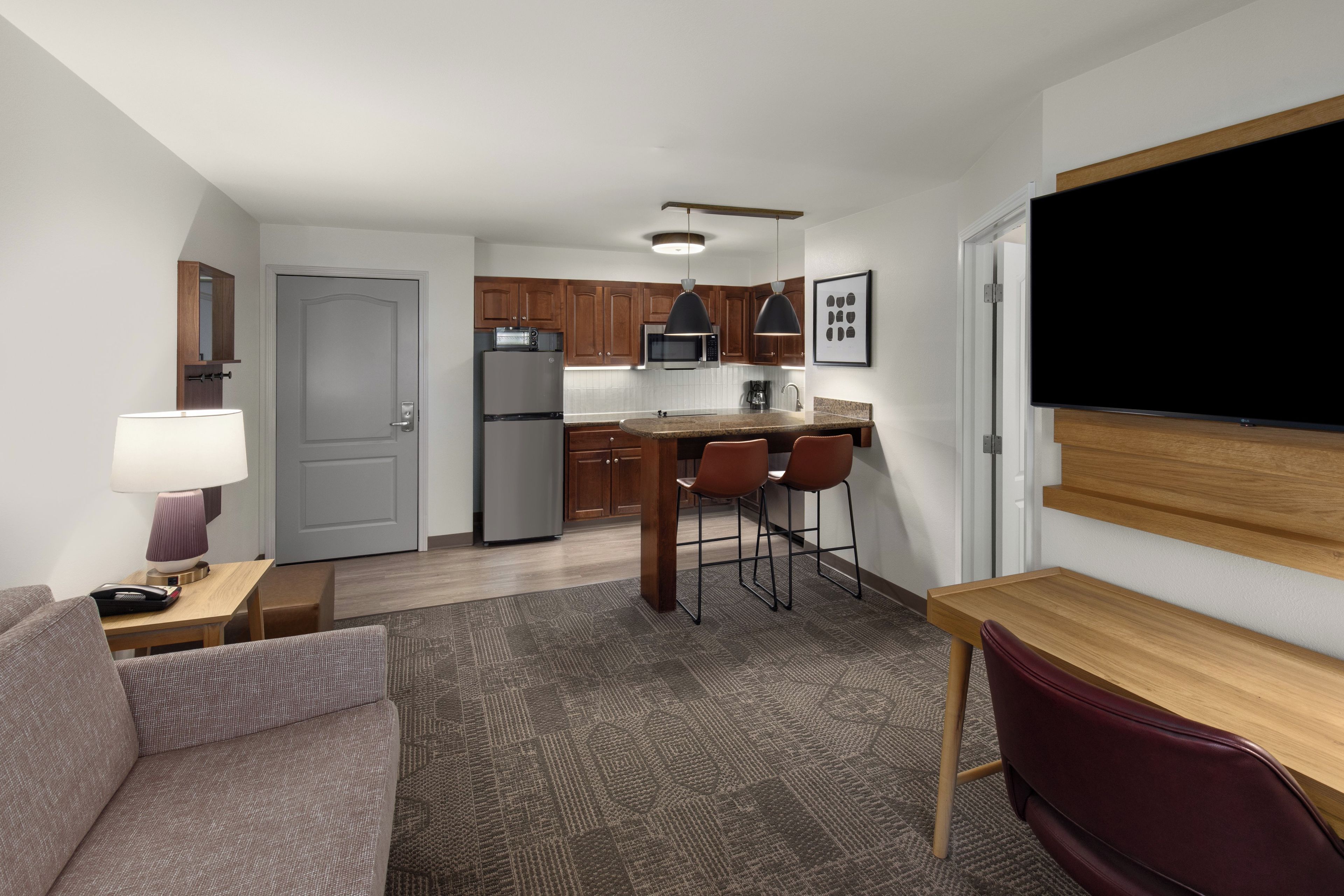 Habitación de hotel compacta con kitchenette, zona de comedor con mesa y sillas, sofá y televisor de pantalla plana montado en la pared.