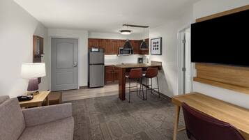 Habitación de hotel compacta con kitchenette, zona de comedor con mesa y sillas, sofá y televisor de pantalla plana montado en la pared.