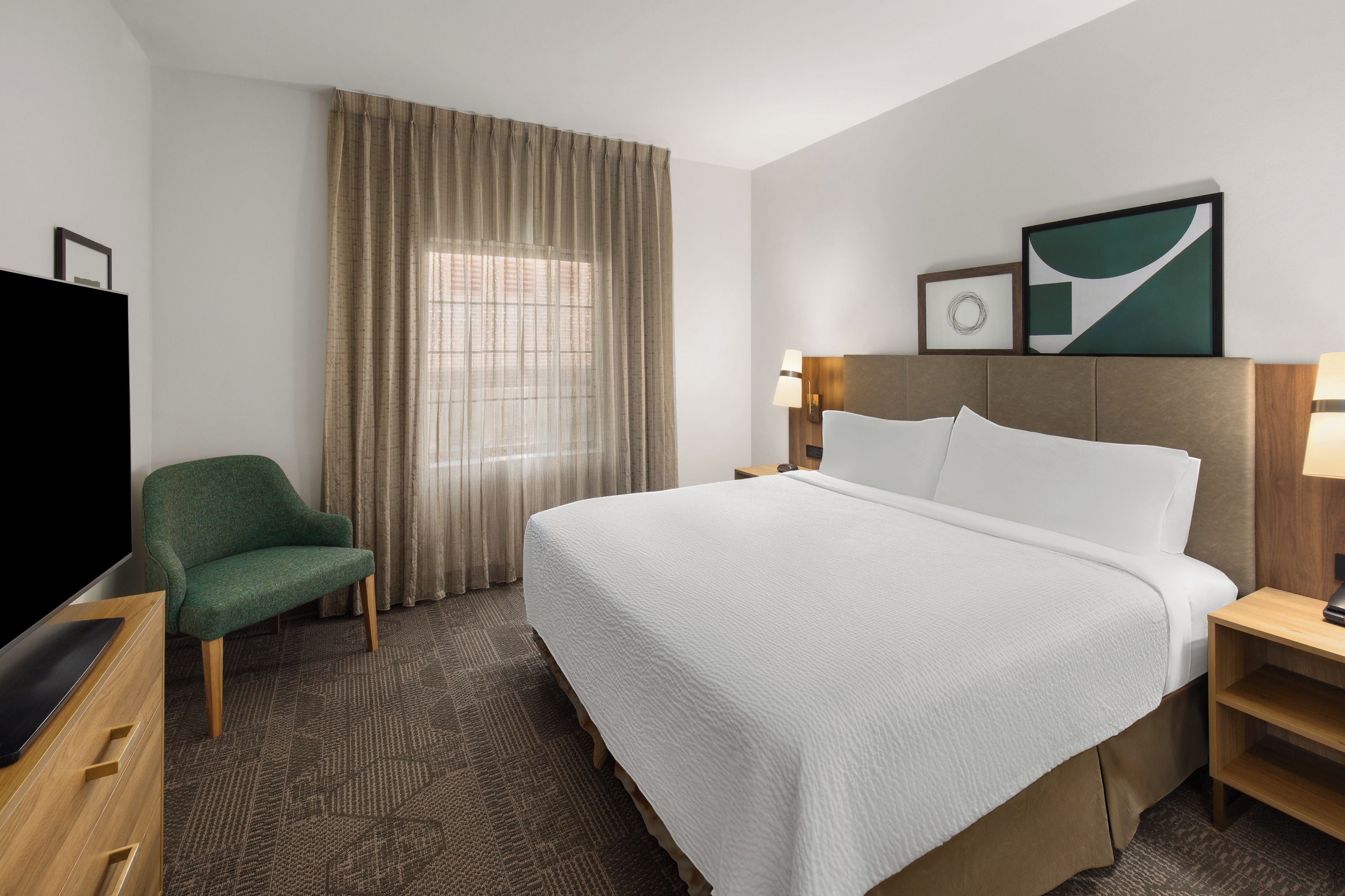 A hotel room with a large bed, a television, a chair, a desk, and a window with curtains.