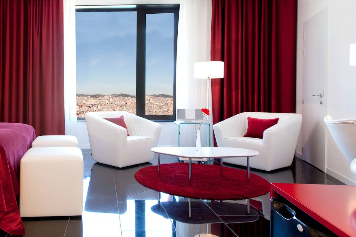 Habitación de hotel con cama, dos sillones blancos, una mesita redonda y vista a un paisaje urbano a través de un ventanal grande.