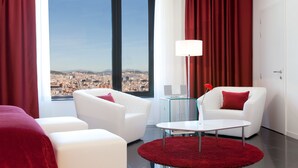 Habitación de hotel moderna con vista a la ciudad, muebles blancos, alfombra roja y una mesa de cristal.