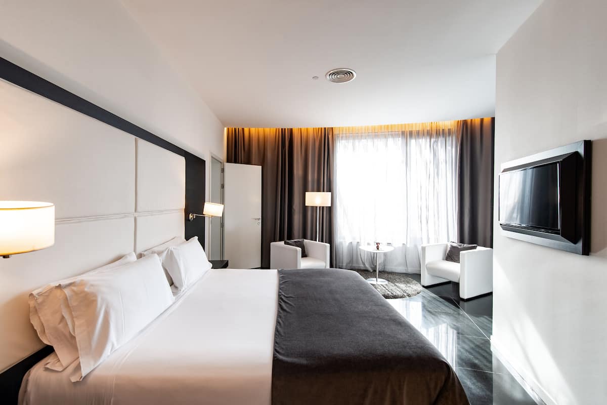 Una habitación de hotel moderna con una cama grande, un televisor montado en la pared, un rincón de descanso con una pequeña mesa y vistas al exterior a través de amplios ventanales.