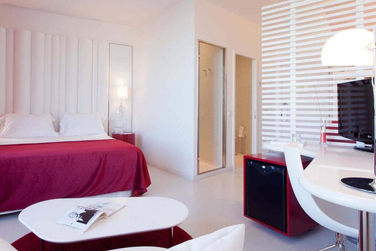 Habitación de hotel moderna con una cama grande, un escritorio, una silla, un televisor y un baño visible a través de una puerta de vidrio.