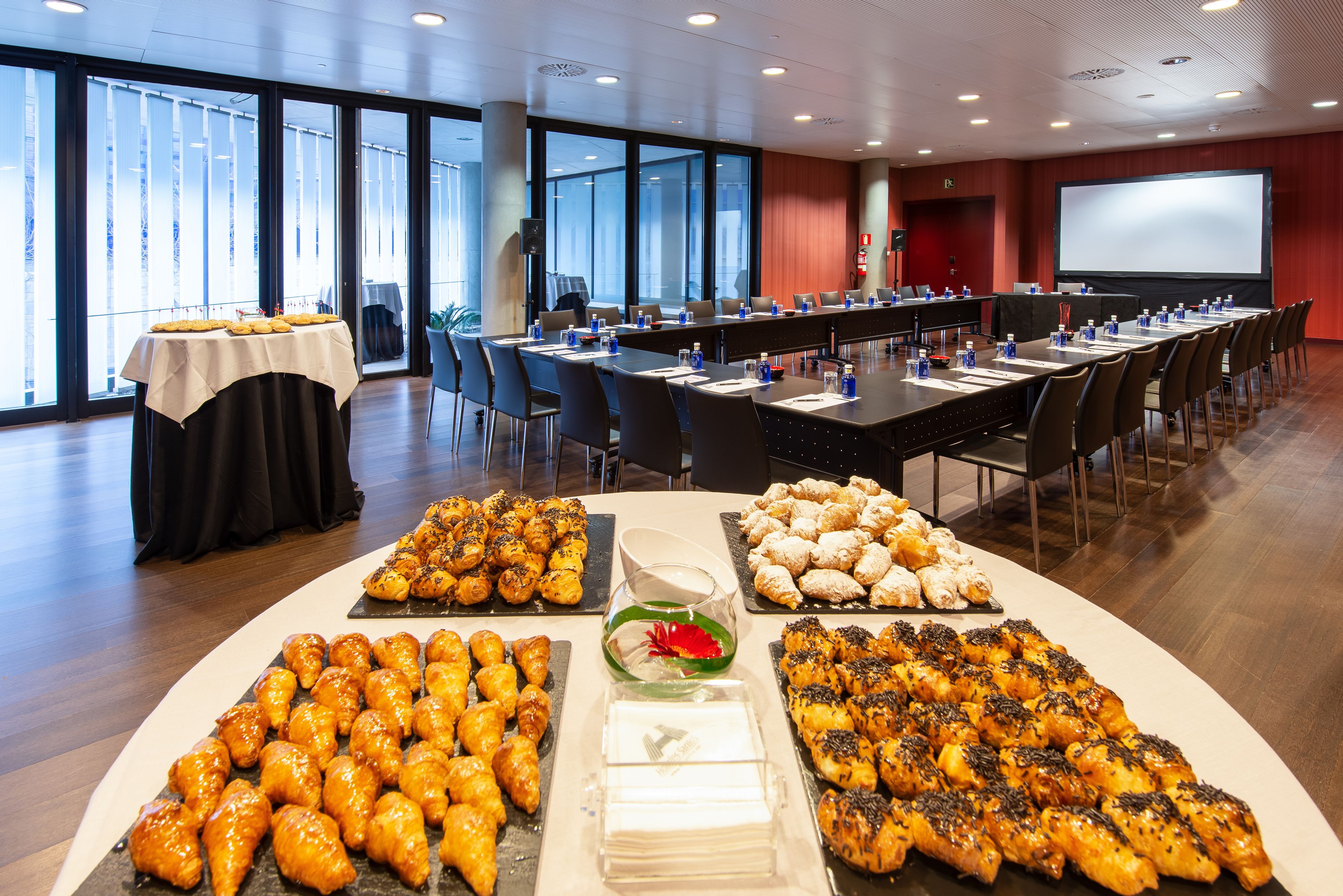 Un salón de conferencias con una larga mesa dispuesta para una reunión, rodeada de sillas y una mesa de buffet con diversos alimentos.