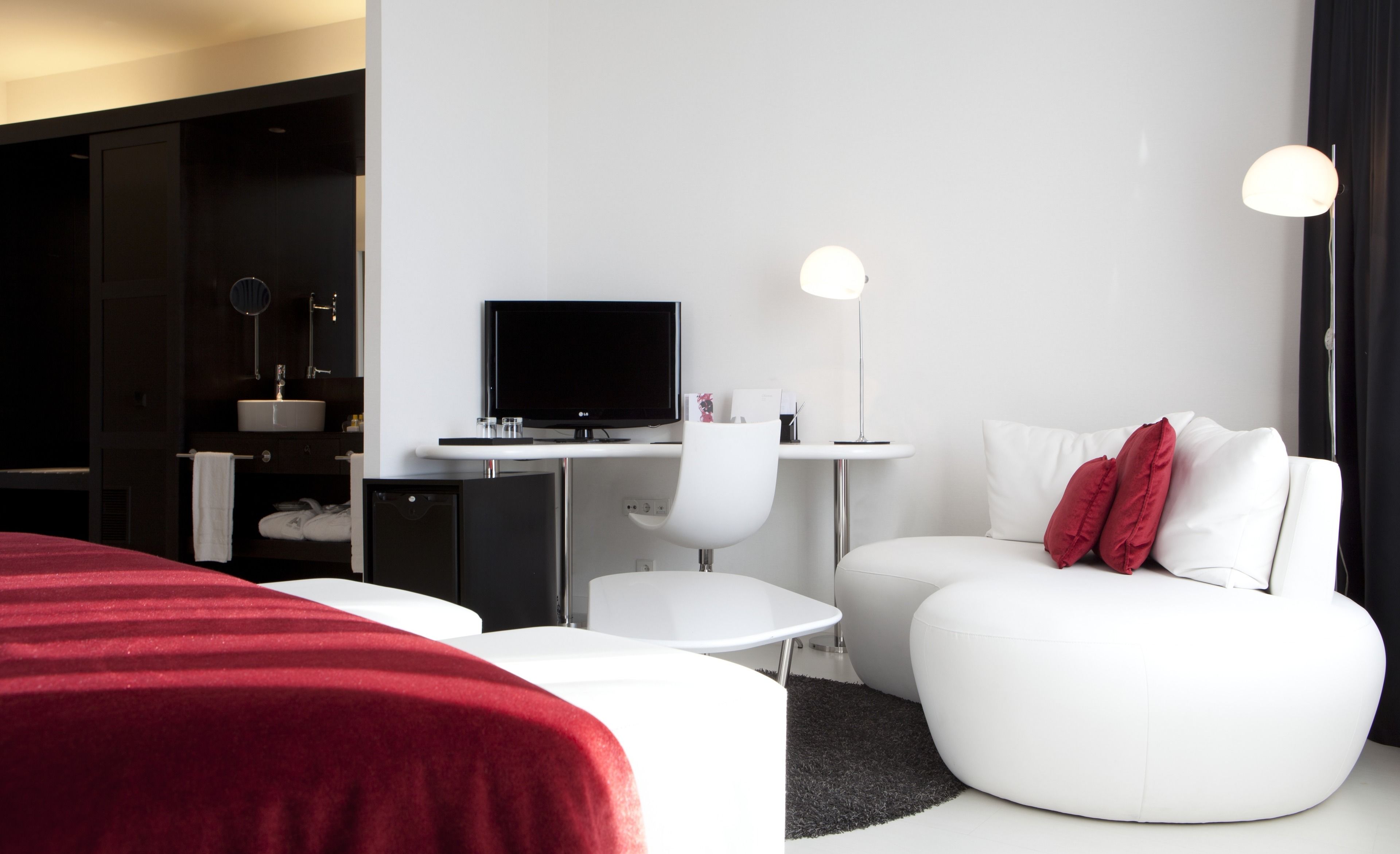 Ein modernes Hotelzimmer mit einem großen Bett, einem Schreibtisch mit Computer, einem weißen Stuhl, einer Couch mit roten Kissen und einem kleinen Tisch.