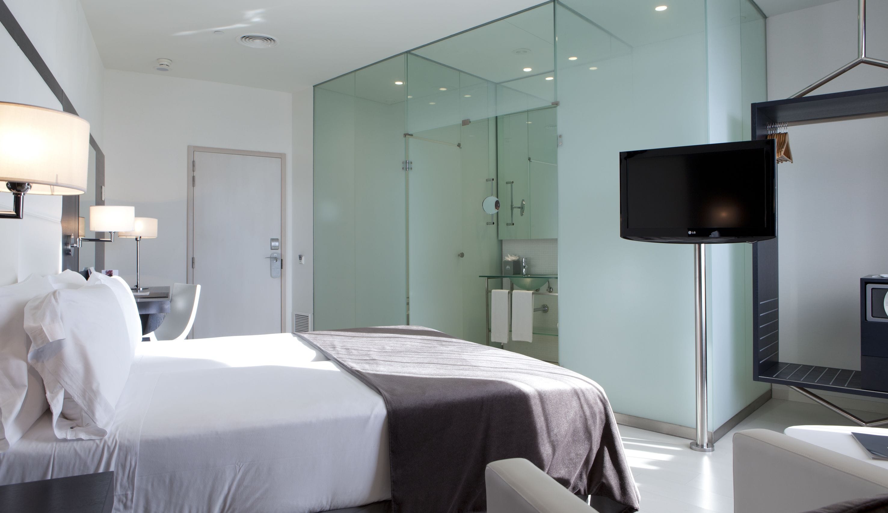 A modern hotel room with a bed, bedside lamps, a TV, and a glass-enclosed shower area.