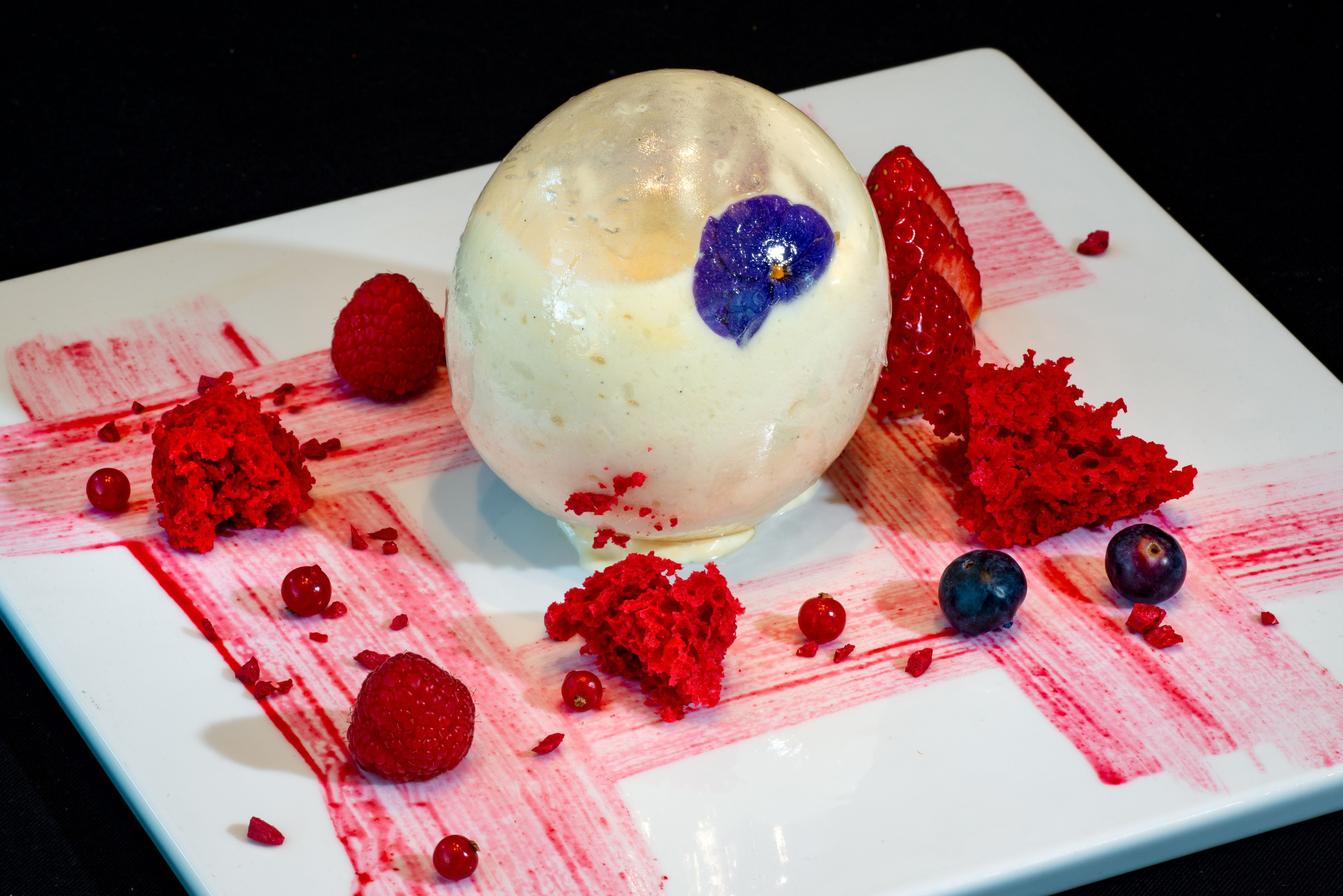 Un postre con base blanca esférica, una rodaja de fresa y frambuesas y arándanos esparcidos.