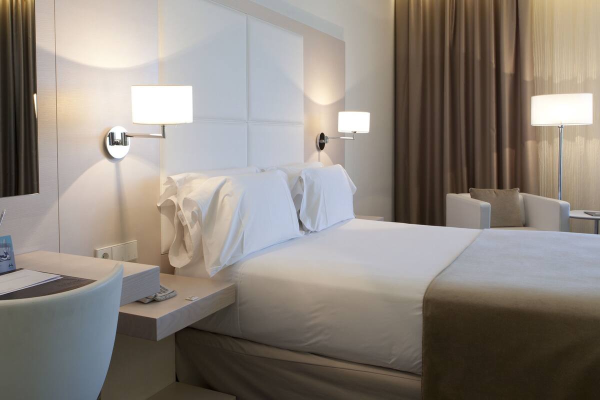 Habitación de hotel con una cama grande, un escritorio con una silla, dos lámparas de noche, un televisor de pantalla plana y una ventana con cortinas.