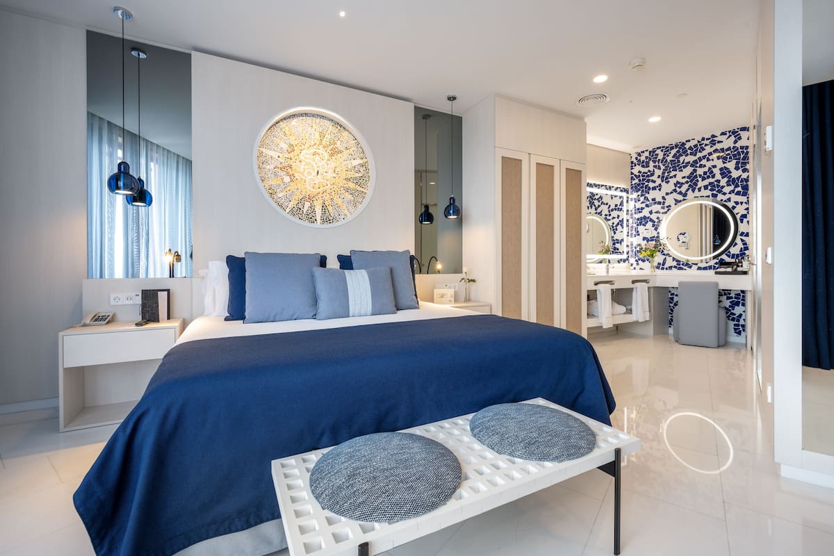 Una habitación de hotel moderna con una cama grande, una mesita de noche, un escritorio, un espejo y un baño con lavamanos y espejo.