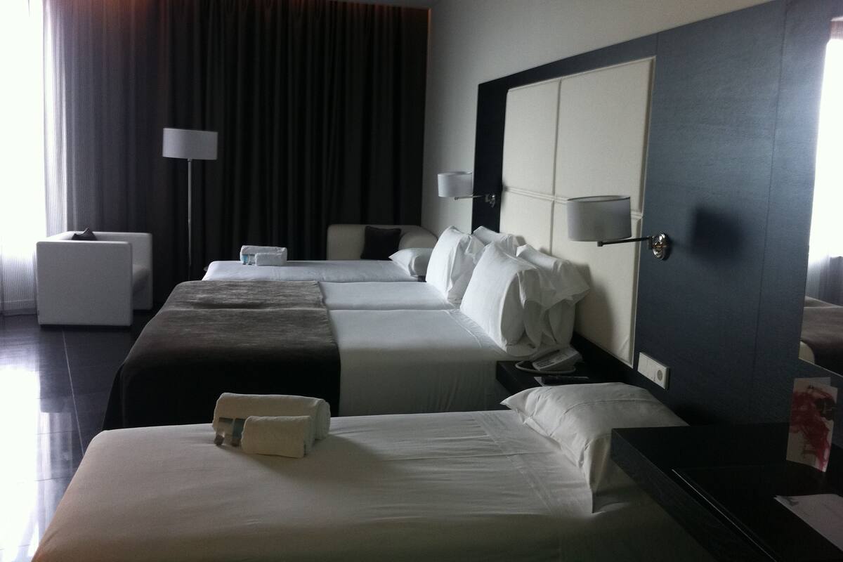 Habitación de hotel con dos camas, una mesita de noche, una lámpara, una silla y un amplio ventanal con cortinas.