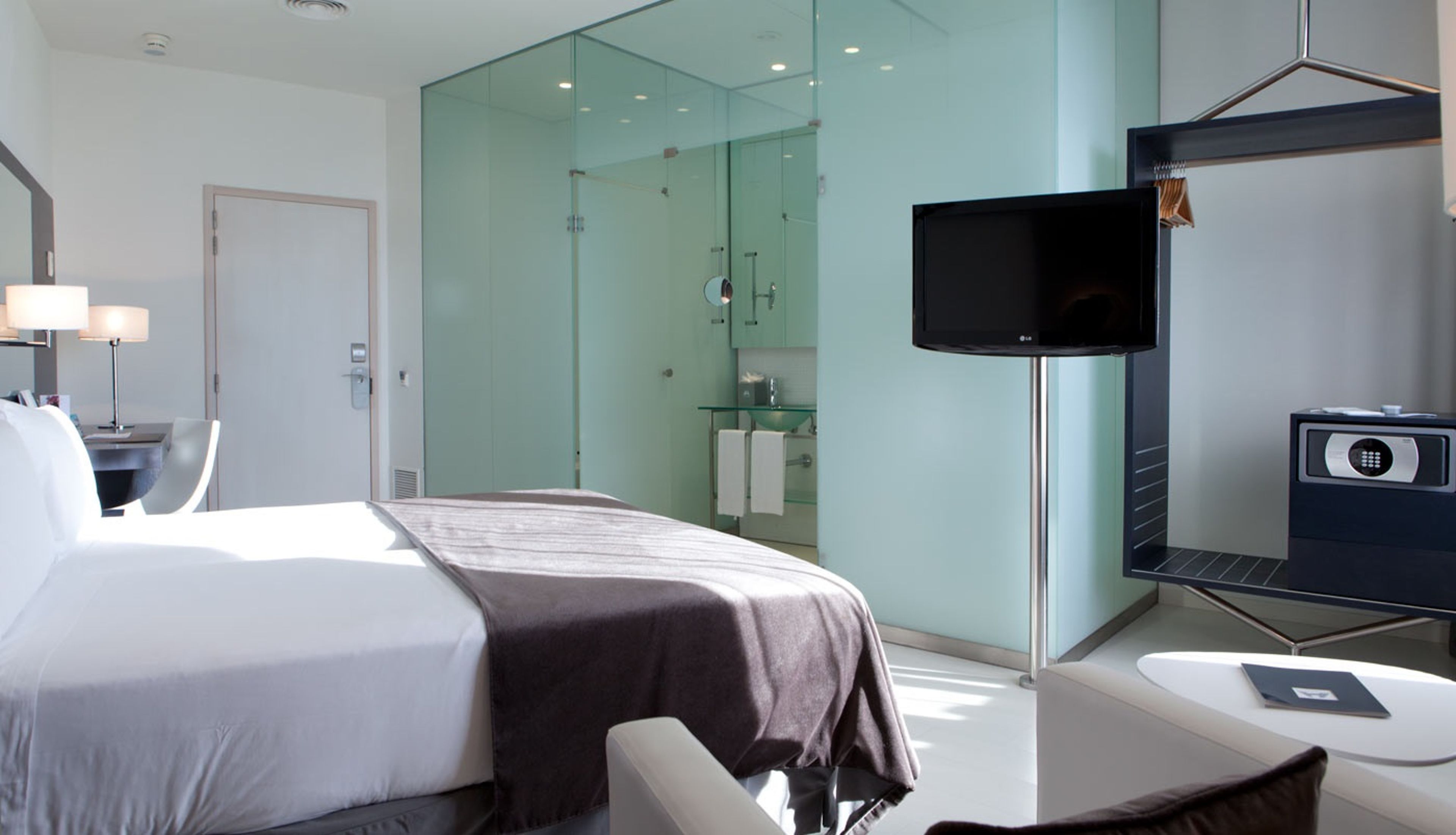 Ein modernes Hotelzimmer mit einem großen Bett, einem Fernseher, einem Schreibtisch mit einer Lampe und einem Glaskastenbad.