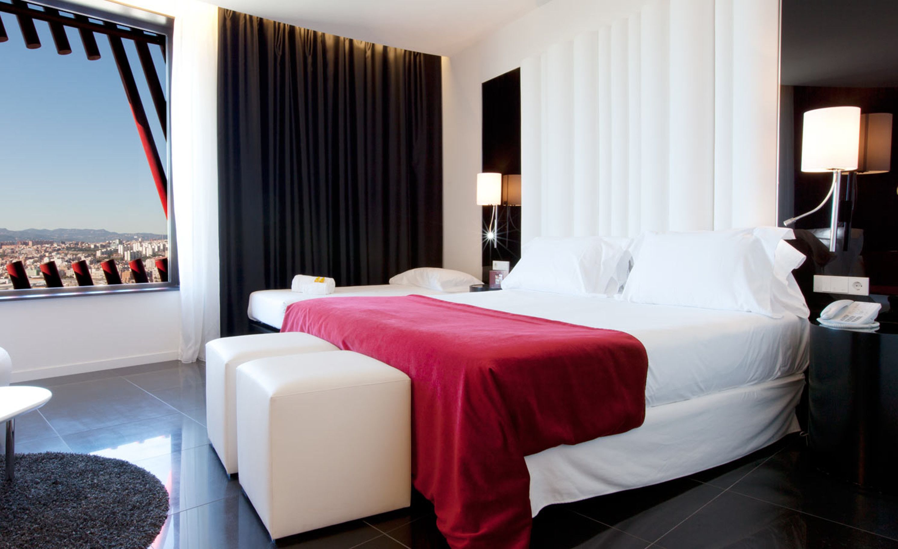 A hotel room with a bed, a desk, a chair, a window with a view of a cityscape, and a nightstand with a lamp.