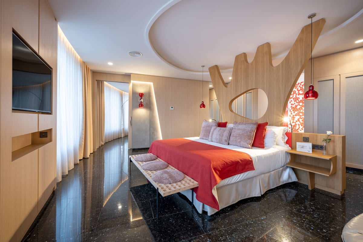 Una habitación de hotel moderna con una cama grande, cabecera de madera, mesita de noche y televisor montado en la pared.