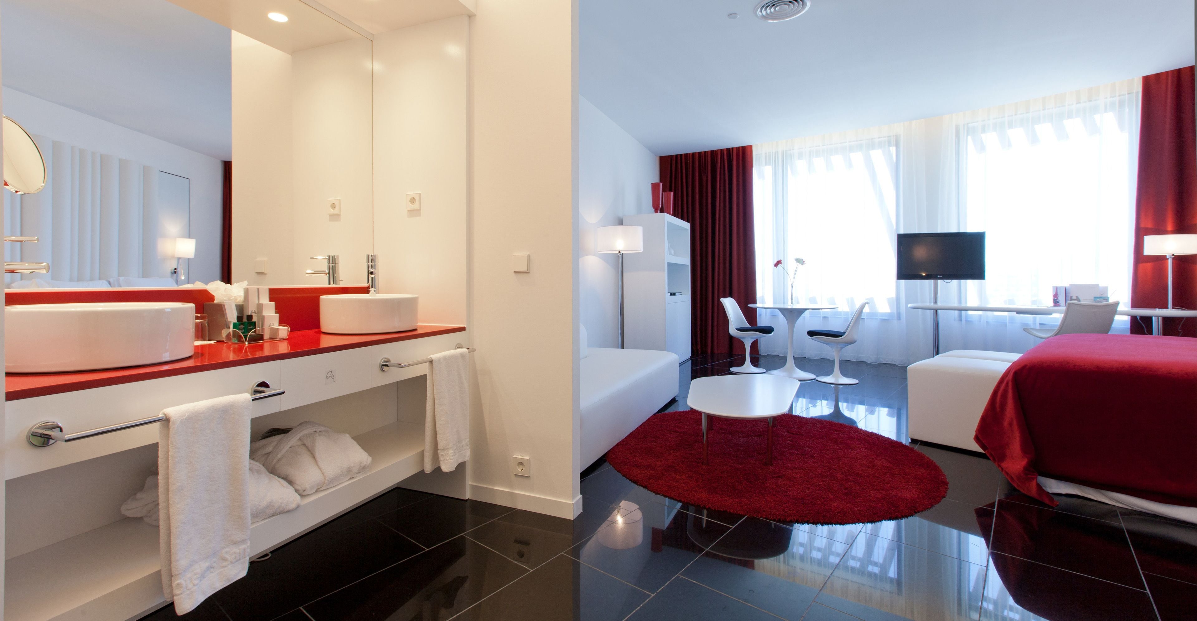 A modern hotel room with a large mirror, a red accent wall, a white sofa, a red rug, and a flat-screen TV.