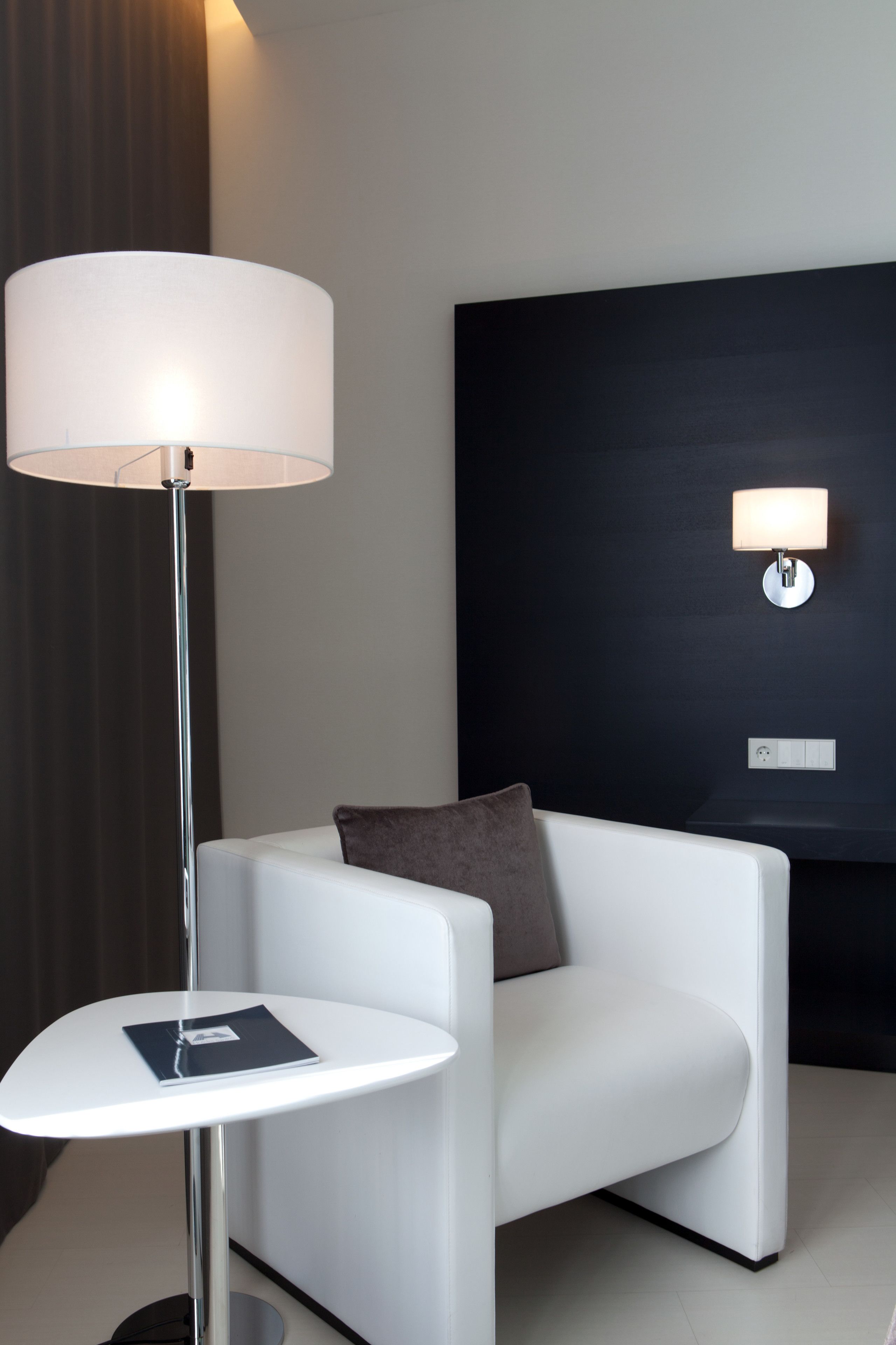 Una habitación de hotel moderna con un sofá blanco, una mesita redonda pequeña, una lámpara de pie y un aplique de pared.