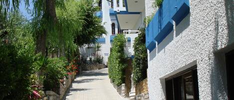 A narrow cobblestone street flanked by buildings with white and blue facades, stone walls, and greenery.