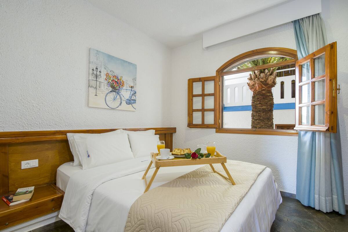 A hotel room with a bed, a tray with breakfast, a painting, and a window with a view.