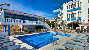A pool area with lounge chairs, a staircase, and a building with a blue and white facade. - Folia Apartments (Chania)