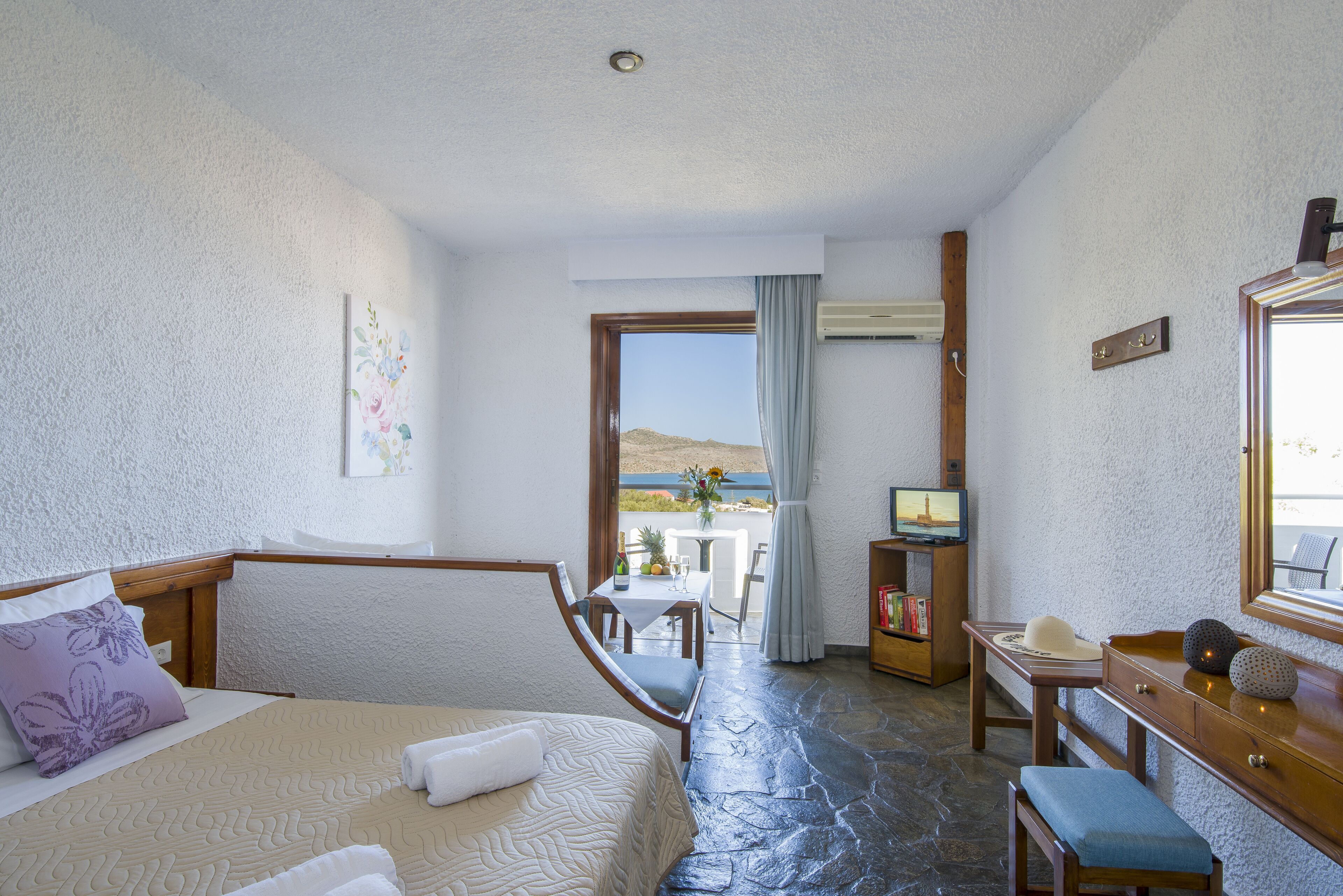 A hotel room with a bed, a desk, a chair, and a small table with a bottle and glasses. There is a window with a view of the outdoors.