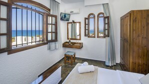 A hotel room with a bed, a wooden wardrobe, a small stool, a mirror, a television, and a window with a view of the sea. - Folia Apartments (Chania)