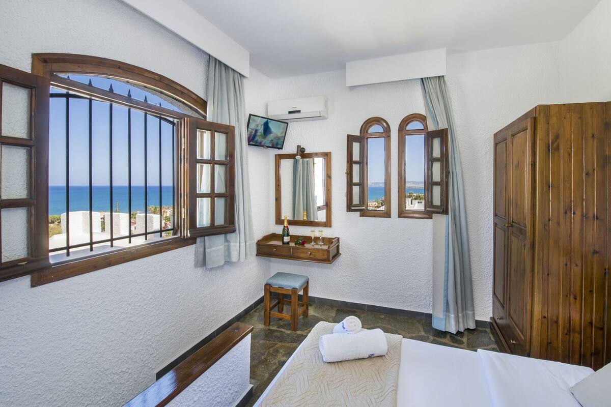 A hotel room with a bed, a wooden wardrobe, a small stool, a mirror, a television, and a window with a view of the sea.