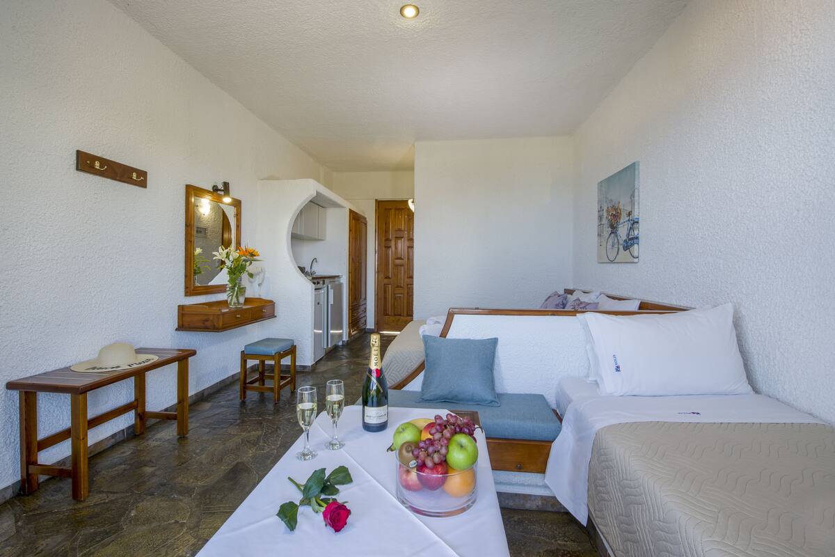 A hotel room with a bed, a small table with a bottle of wine, glasses, and a bowl of fruit. There is a mirror on the wall and a painting above the bed.