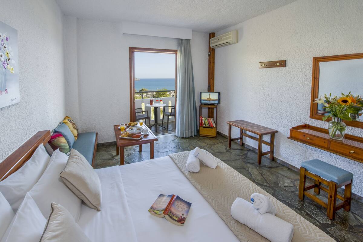 A hotel room with a bed, a small table with a book, a TV, a balcony with a view of the sea, and a mirror with flowers.