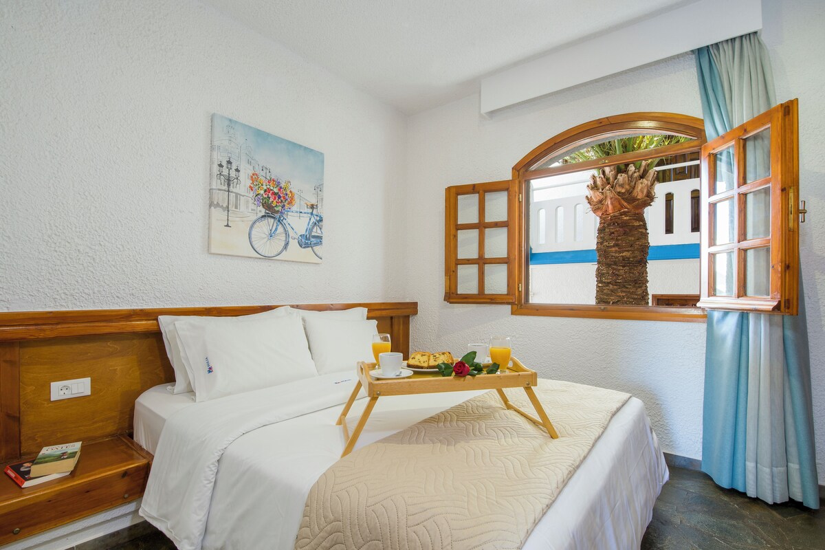A hotel room with a bed, a tray with breakfast, a painting, and a window with a view.