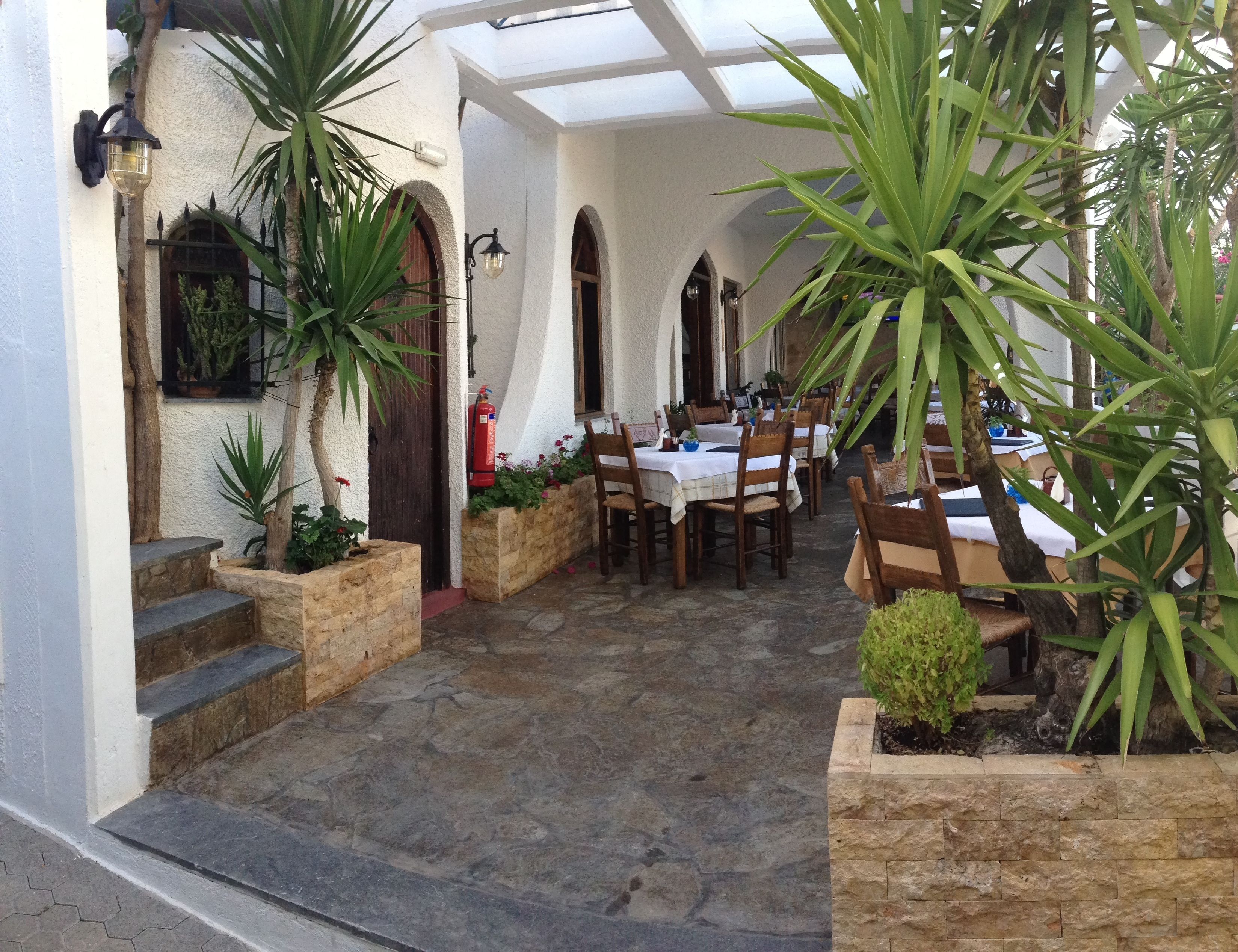 A covered outdoor dining area with stone steps leading up to a white building, featuring arched doorways and potted plants.