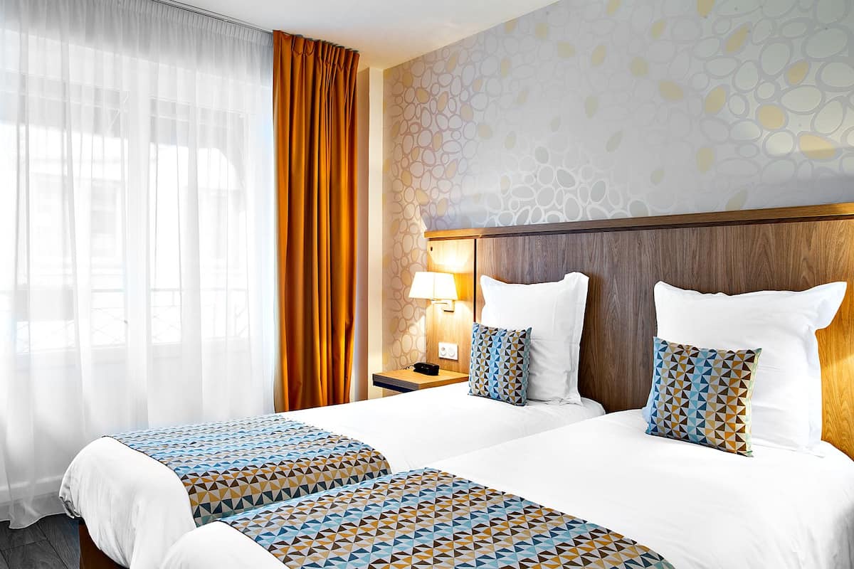 Une chambre d’hôtel avec deux lits, une table de chevet avec une lampe, un mur décoré d’un motif et une fenêtre avec des rideaux légers.