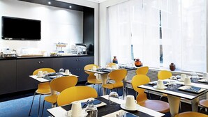 Un espace repas moderne dans un hôtel, avec des chaises jaunes, des tables noires et un téléviseur à écran plat fixé au mur.