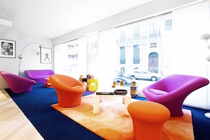 A modern living room with vibrant purple and orange chairs, a wooden coffee table, and a view of a city street through large windows. - Logis Hôtel La Caravelle (Toulouse)