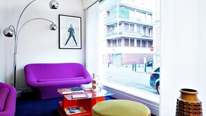 Un salon moderne avec un canapé violet, une table basse rouge et un pouf jaune. Un tableau encadré est accroché au mur, et une lampe sur pied se trouve dans un coin de la pièce.
