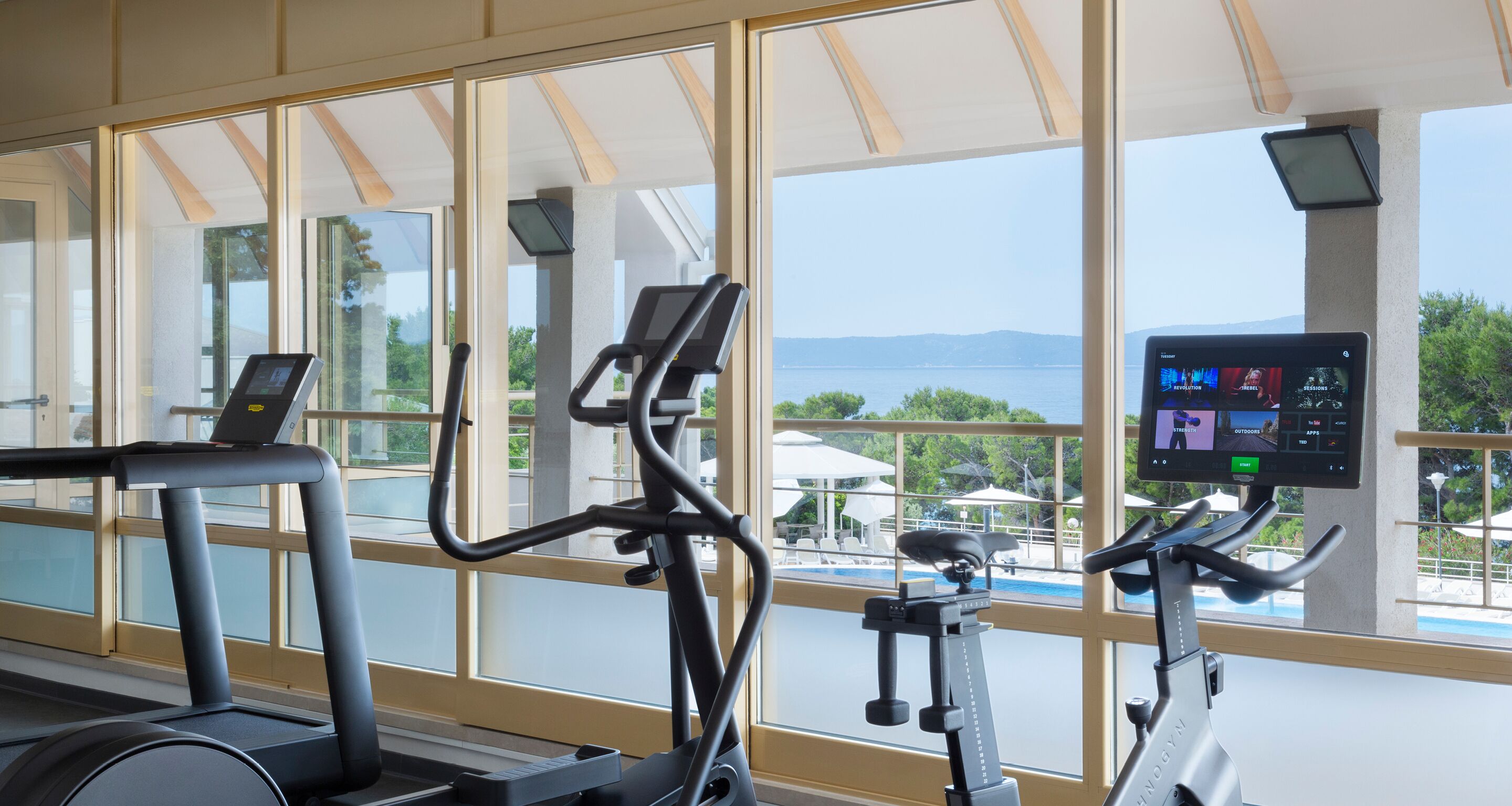A gym with exercise equipment and a large window overlooking a pool and mountains.