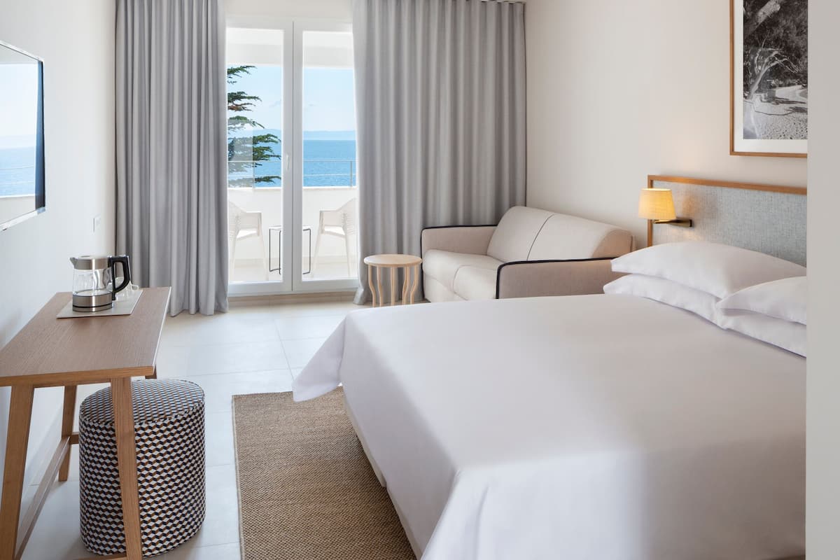 Habitación de hotel con cama, escritorio, silla y vista al mar a través de una puerta corrediza.