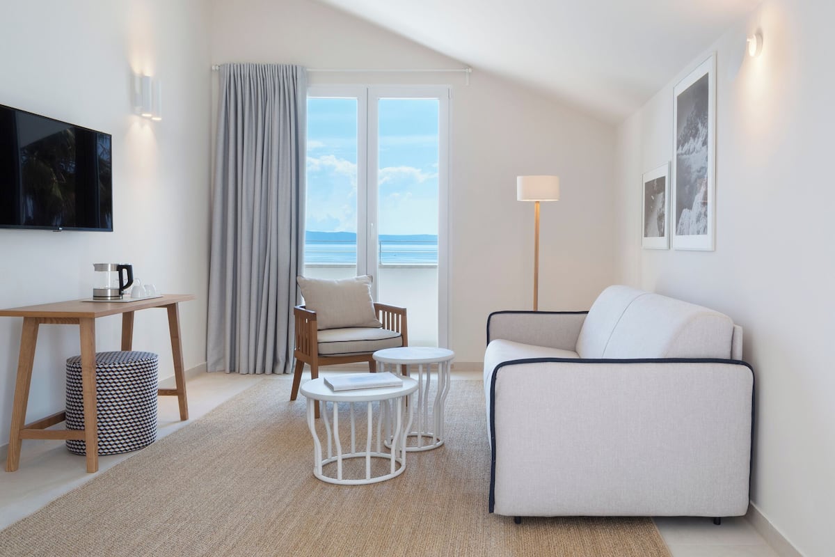 Habitación de hotel moderna con sofá, sillón, mesitas, televisor y vista al mar a través de ventanas amplias.