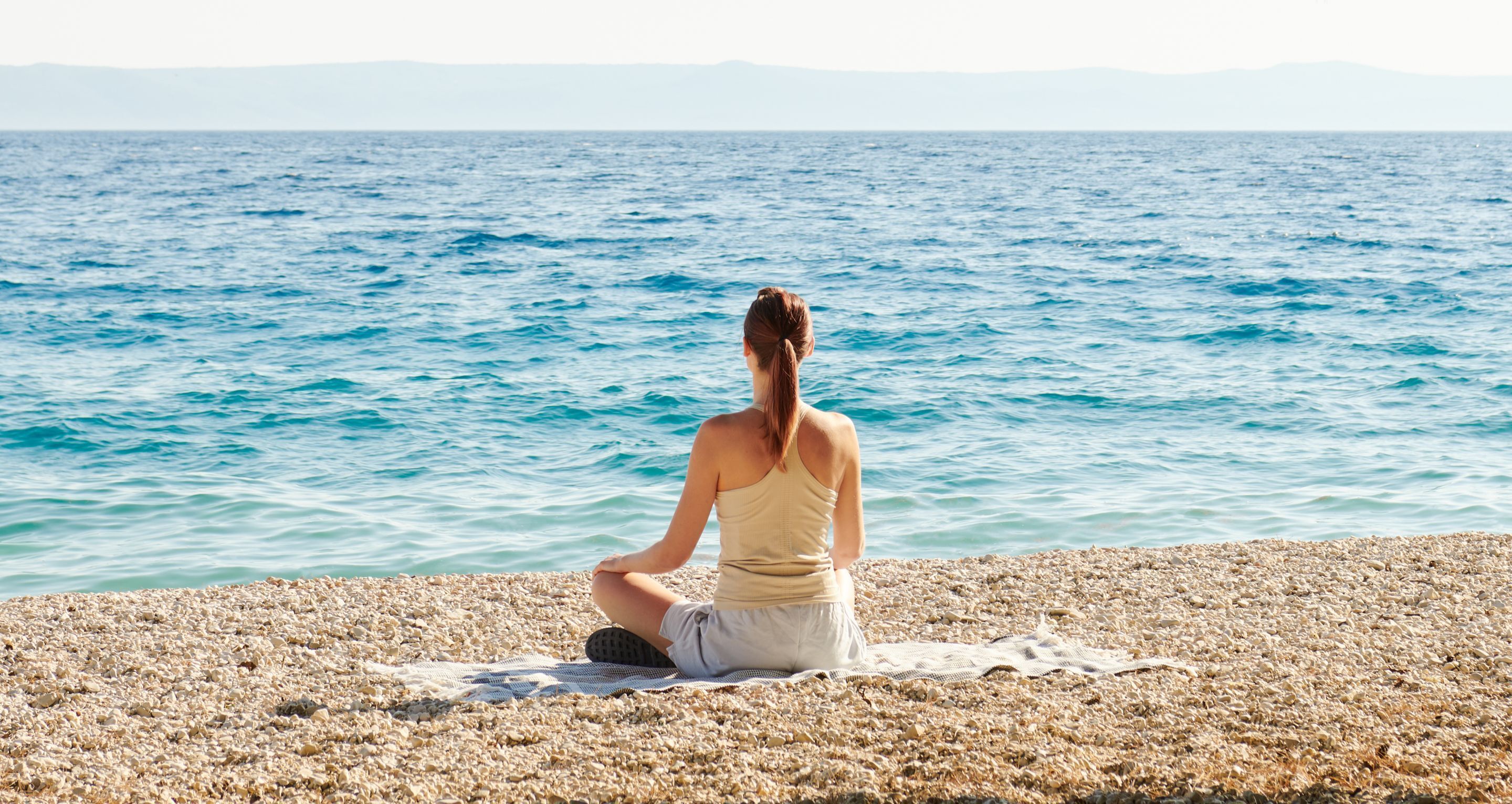 Una persona meditando en una playa de guijarros, con vista al mar y montañas distantes.