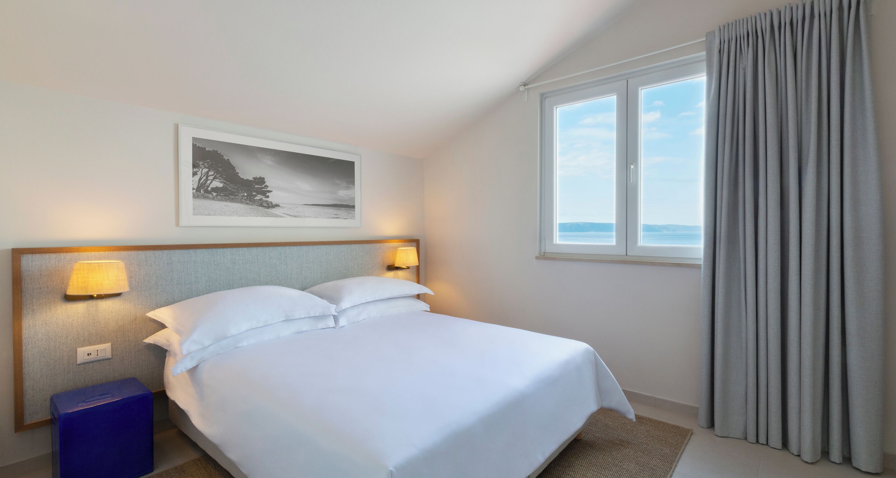 A hotel room with a bed, two bedside lamps, a nightstand, a framed picture on the wall, and a window with curtains.